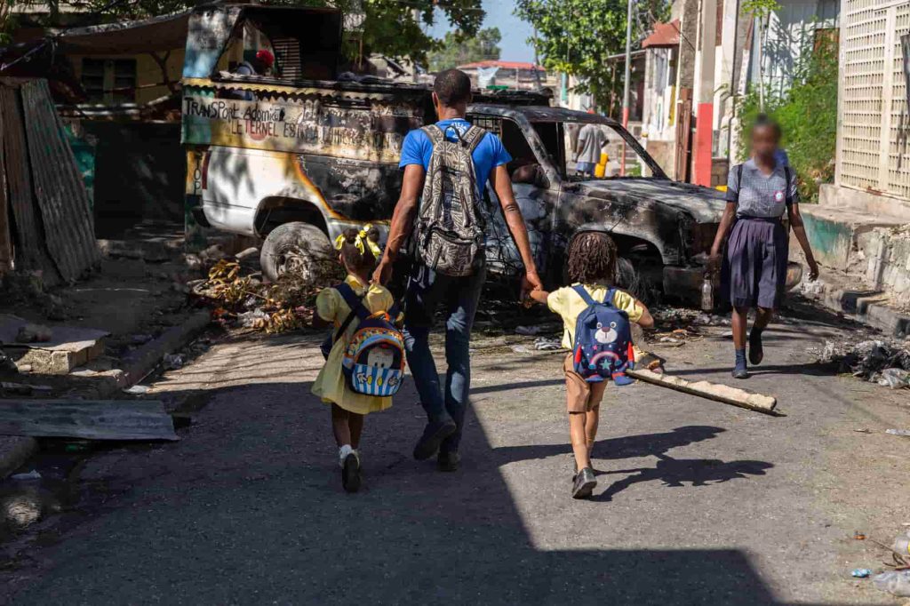 Um homem e duas crianças passam por um carro incendiado por gangues armados na área de Poste Marchand, na capital haitiana. Foto de Guerinault Louis/Anadolu via Getty Images.